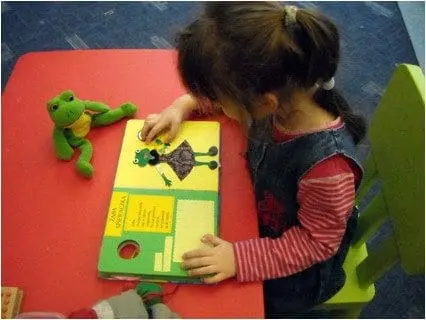 Child at a table examines a touch book with a frog toy nearby.