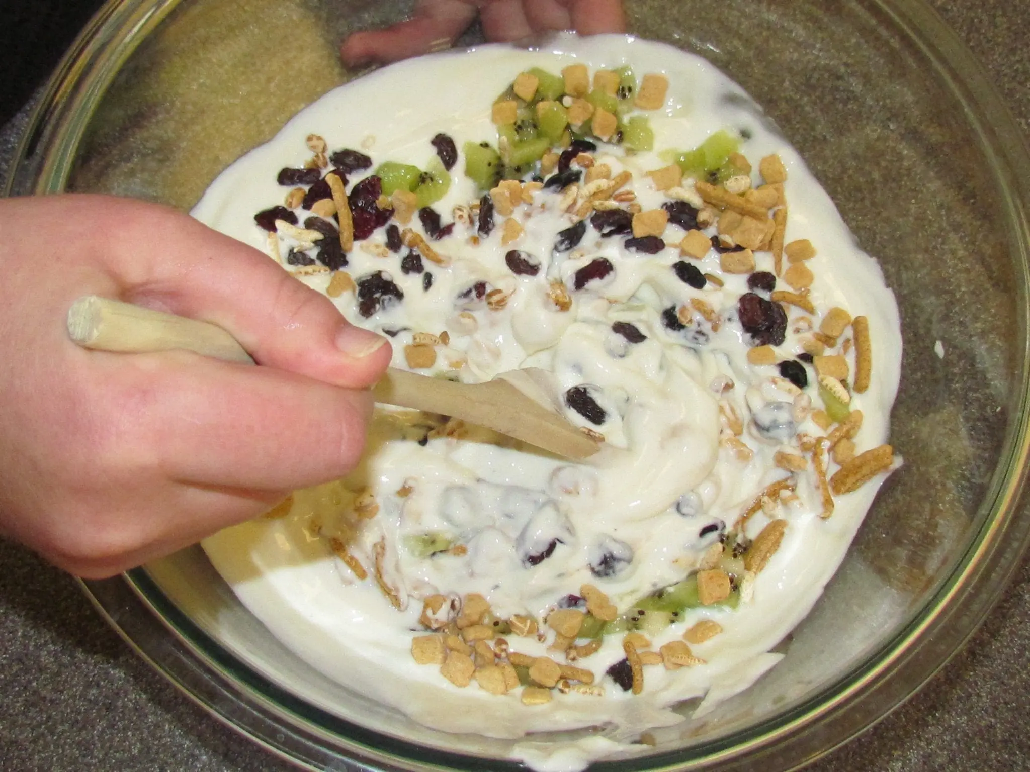 A hand stirs yogurt mixture with dried fruit and granola in a glass bowl.
