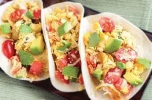 Three breakfast tacos with scrambled eggs, avocado, and tomatoes on a dark platter.