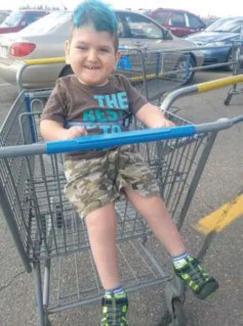 A young child with bright blue hair sits in a shopping cart, smiling.