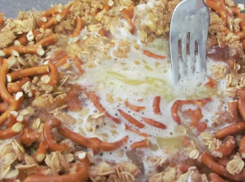 A fork mixes pretzel pieces, granola, melted butter, and brown sugar in a pan.
