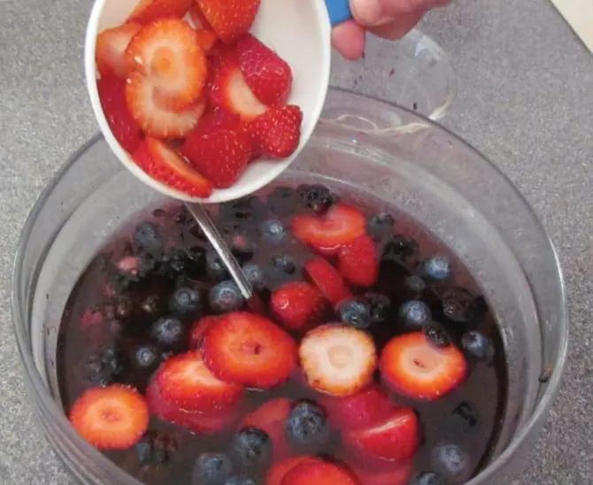 Cup of sliced strawberries being poured into a bowl of gelatin and berries.
