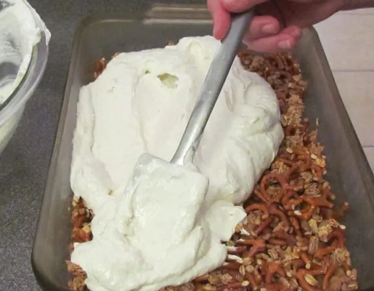 Spoon spreading whipped topping over pretzel and granola base in a glass baking dish.