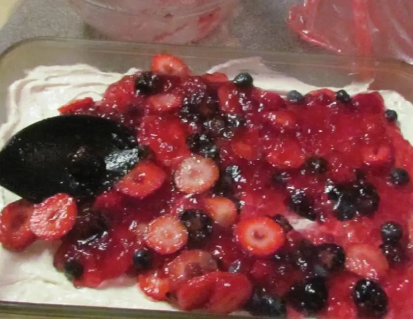 A glass pan with a white creamy base topped with red berries and blueberries, and a black spoon.