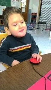 A young boy in a striped shirt uses a red adaptive switch.