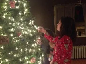 A child in pajamas decorates a Christmas tree with lights and ornaments.