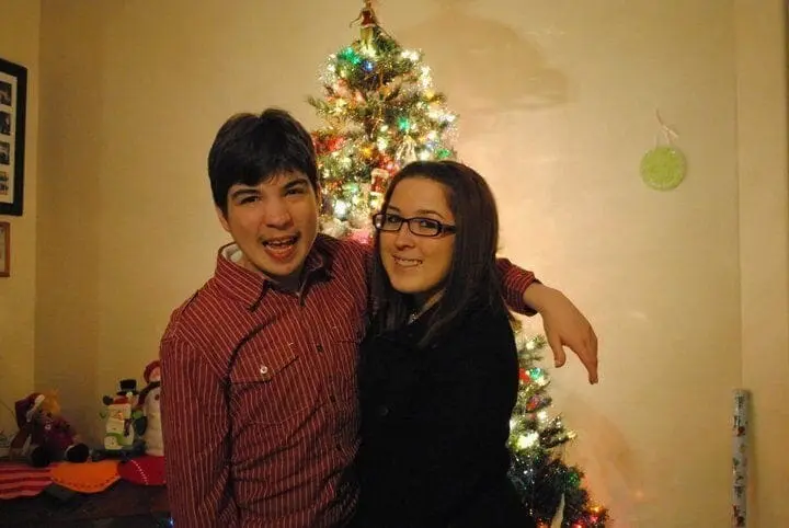 A young man and woman smile in front of a decorated Christmas tree.