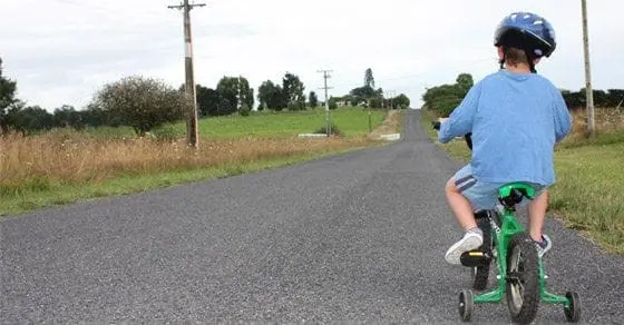 Child wearing a blue shirt and helmet rides a green bicycle with training wheels down a paved road.