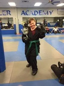 Trey, wearing a karate uniform and green belt, proudly holds a trophy in a gymnasium.