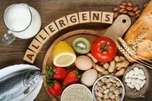 Various foods commonly associated with allergies, including fish, strawberries, eggs, nuts, milk, and bread, arranged around wooden alphabet blocks spelling "ALLERGENS.