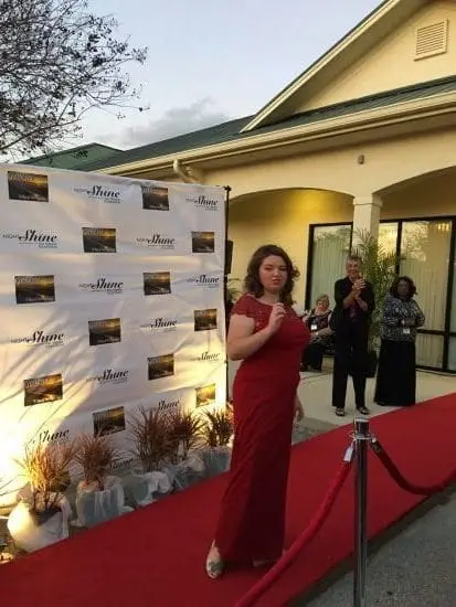 Woman in a red dress walks on a red carpet in front of a step and repeat banner.