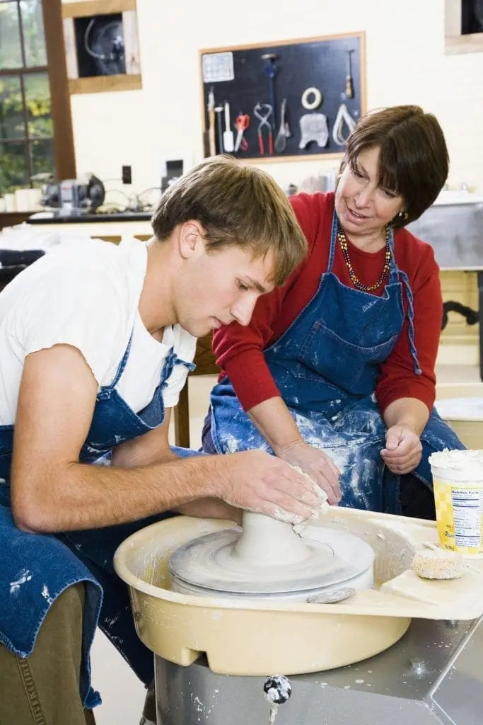 A woman instructs a man as he works on a pottery wheel.