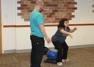 A man in a teal shirt and dark pants observes a girl in a dark shirt and jeans doing a squat.