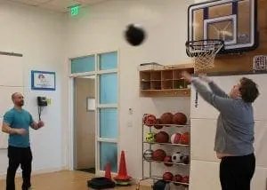 A coach watches as an athlete practices shooting a ball into a basketball hoop mounted on a wall.