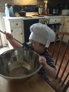 Child in chef hat tasting from a large bowl with a spoon.