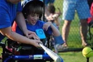 Boy in a wheelchair swings a baseball bat with a coach behind him.