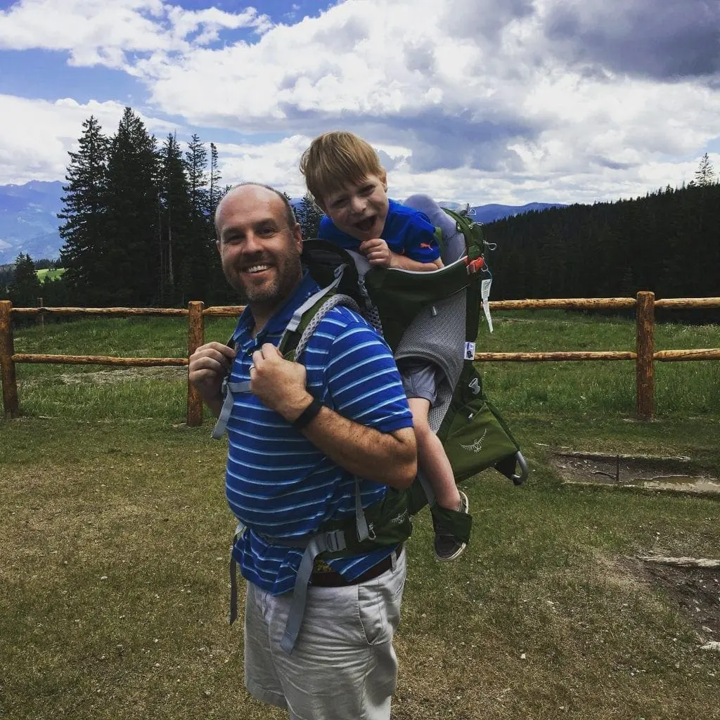 Man carrying a child in a hiking backpack on a trail.