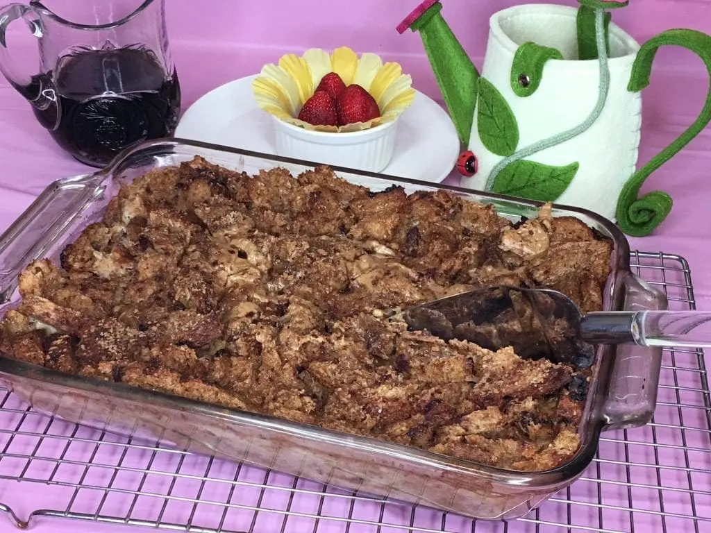Freshly baked French toast casserole in a glass dish with a decorative watering can and a small bowl of strawberries.