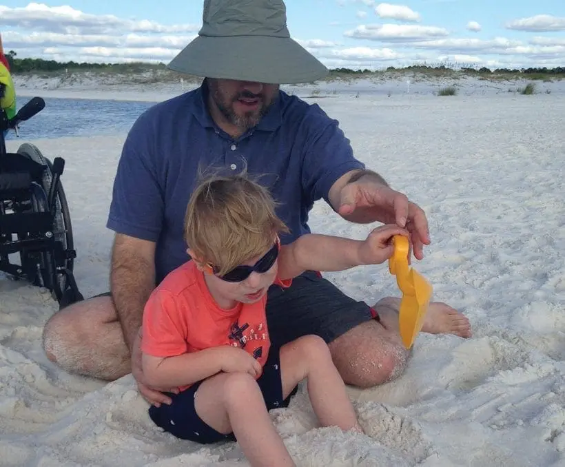 A man in a hat and a child with sunglasses sit on a beach with a toy.