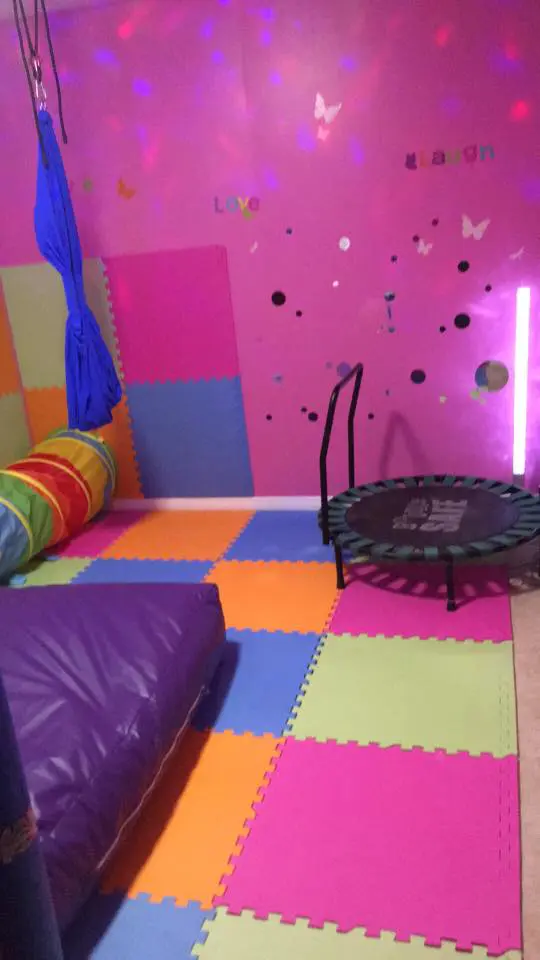 A colorful sensory room with a trampoline, swing, and padded floor.