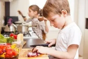 Boy slicing red onion on a cutting board in a kitchen with a girl in the background.