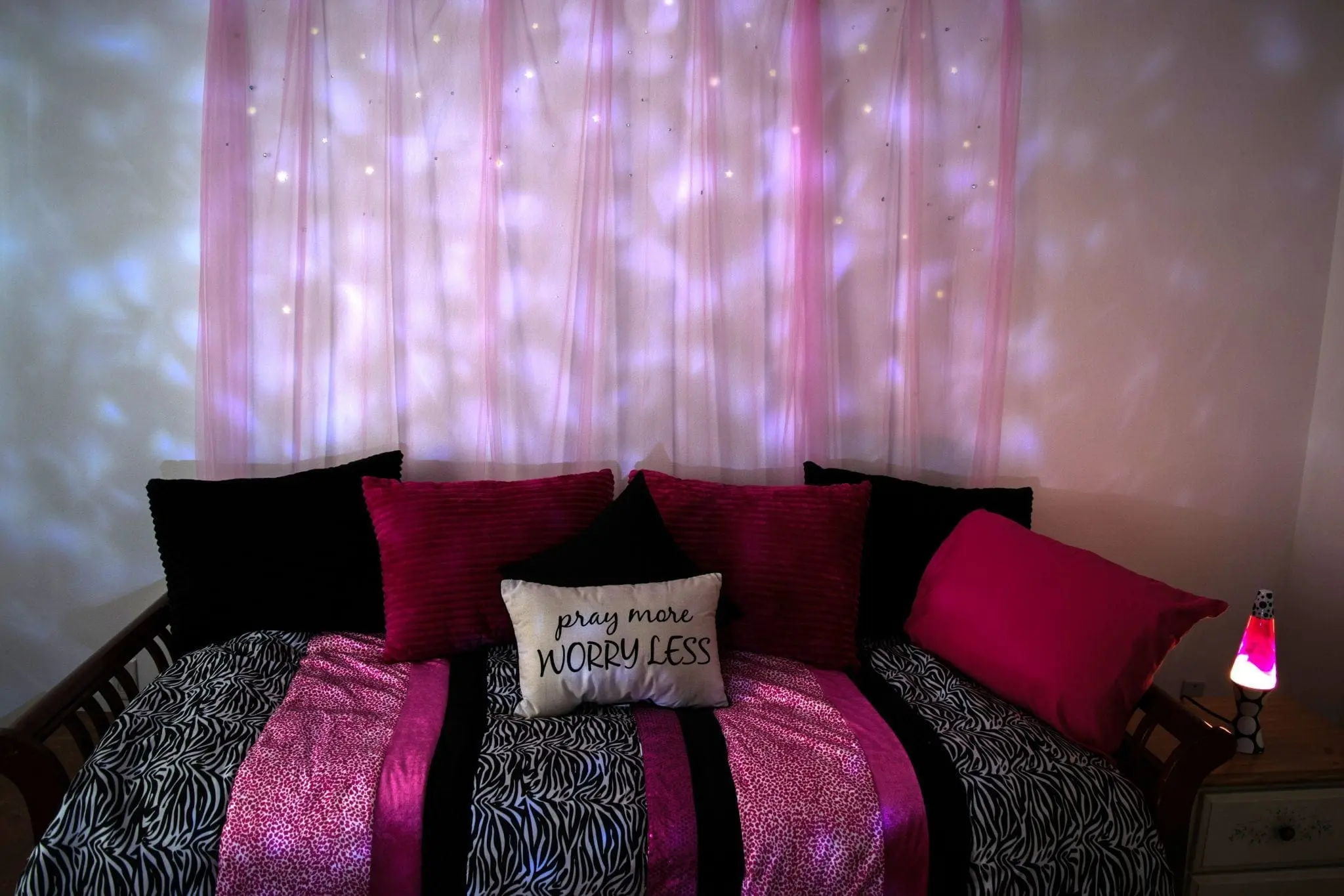 A bed with black, pink, and white pillows, and a bedspread with pink and black zebra stripes, is set against a wall with pink fabric and star-like lights.