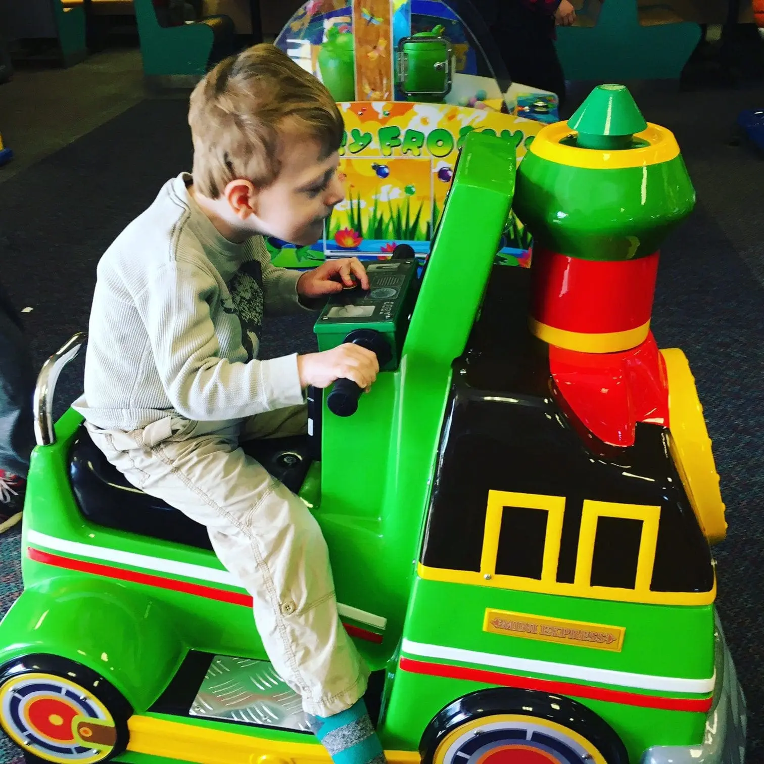Child in a light blue shirt and tan pants riding a green toy train.