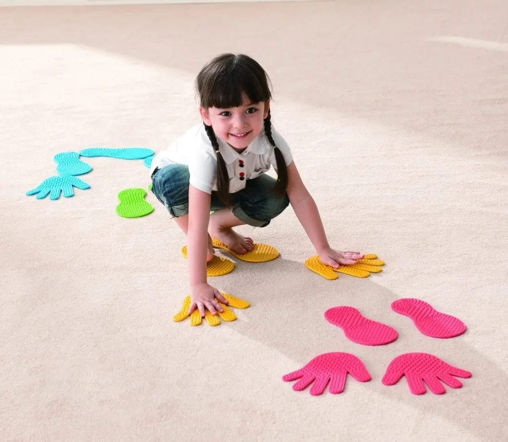 A young girl crouches on a carpeted floor, reaching for colorful stepping stones shaped like hands and feet.