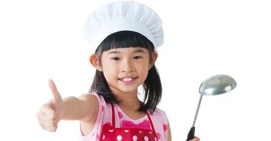 Child in chef hat and apron gives a thumbs up while holding a ladle.