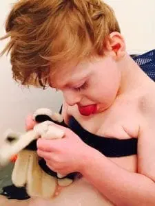A young boy with reddish-blond hair looks down and plays with a white washcloth in a bathtub.