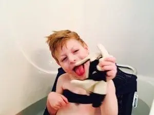 Child in a bathtub holding a stuffed toy with his tongue out