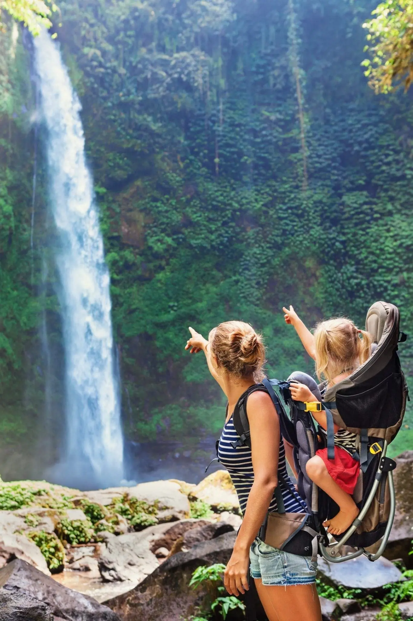 Woman carrying child in backpack points at a waterfall.