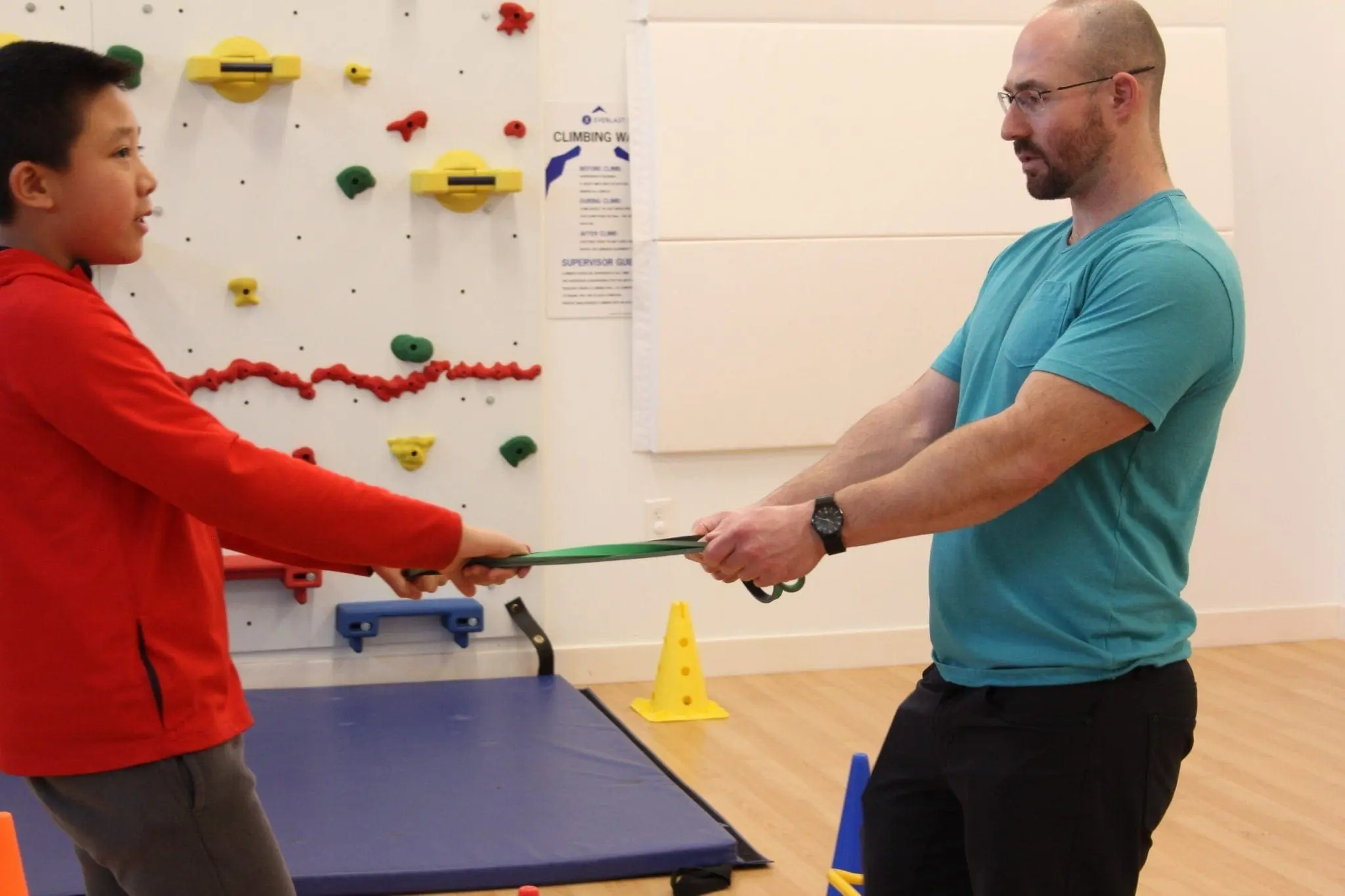 A boy in a red sweatshirt and an adult man in a teal shirt pull on a resistance band.