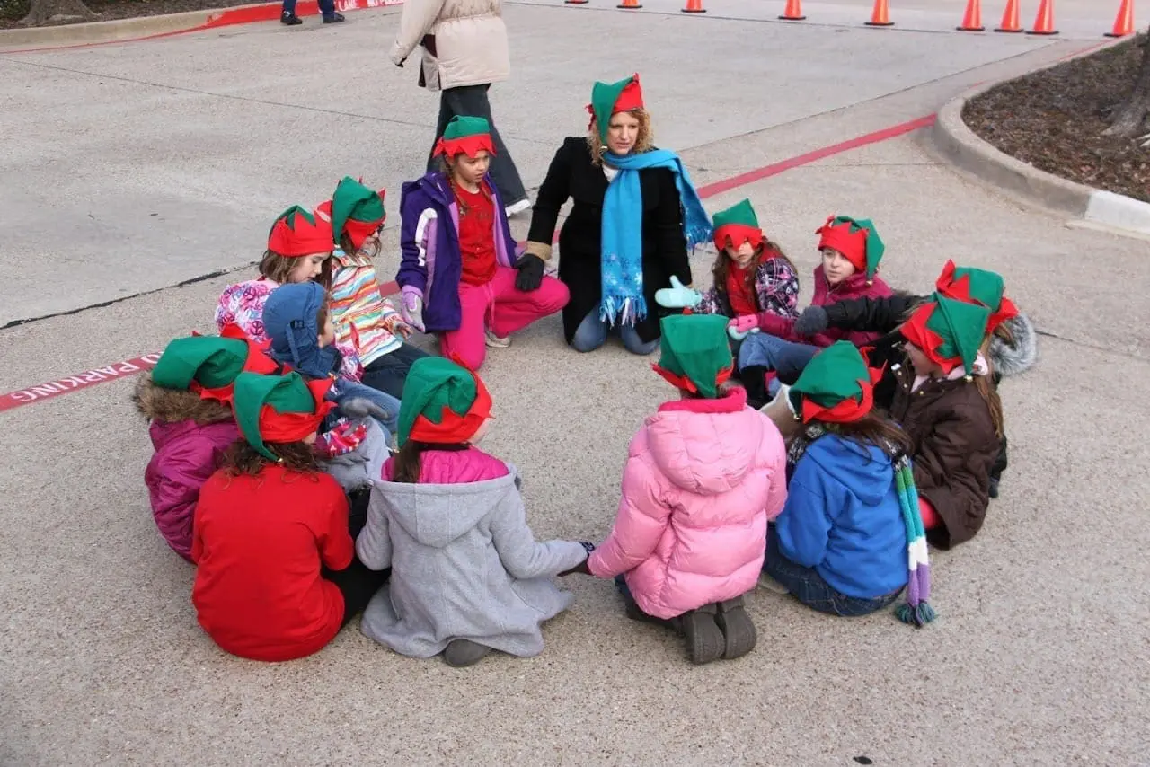 Children wearing elf hats sit in a circle outdoors.