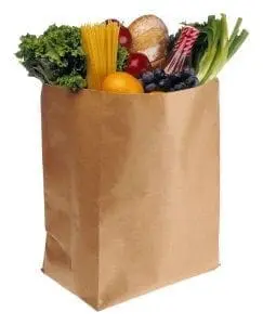 A brown paper bag filled with groceries including spaghetti, kale, grapes, and a loaf of bread.