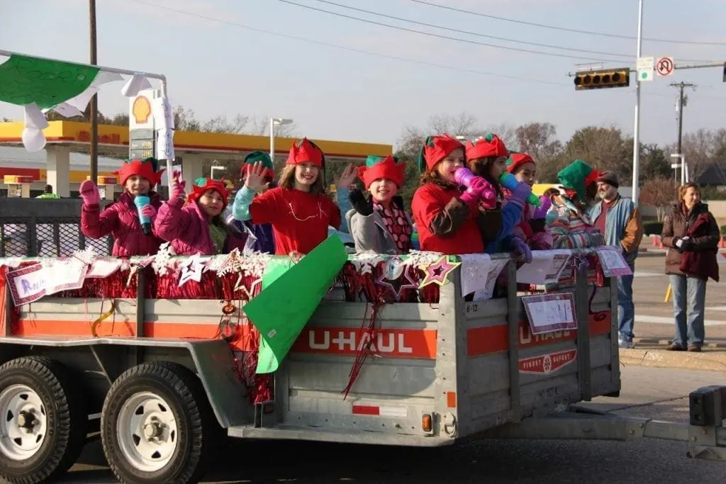 Children in red hats wave from a U-Haul trailer in a parade.
