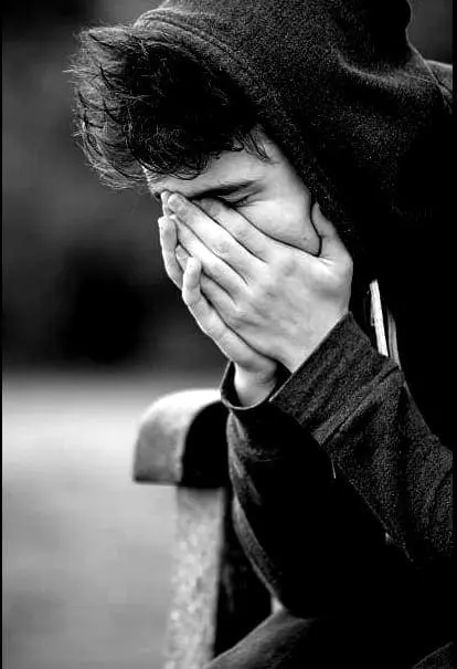 Young person in a hooded sweatshirt covering their face with their hands.