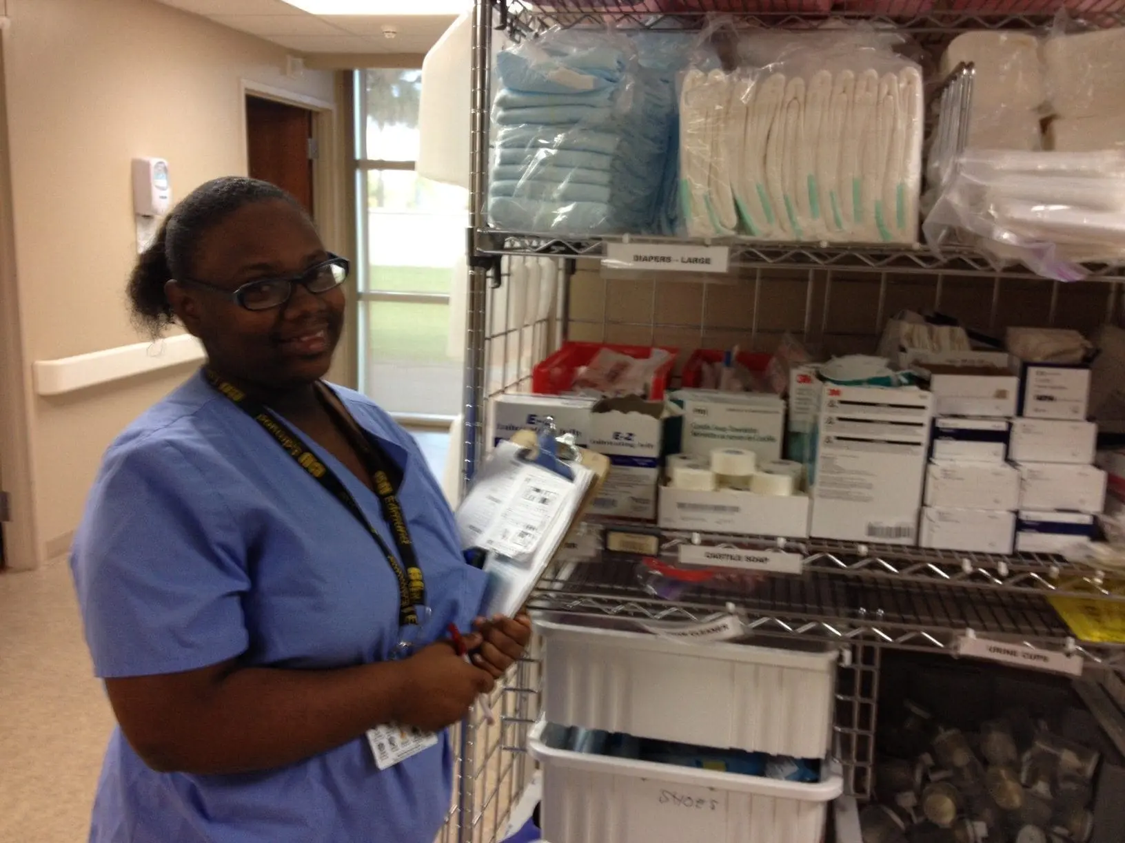 A woman in blue scrubs holds a clipboard next to a shelf of medical supplies.