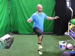 A man in a blue t-shirt and black shorts performs a single-leg squat on artificial turf.