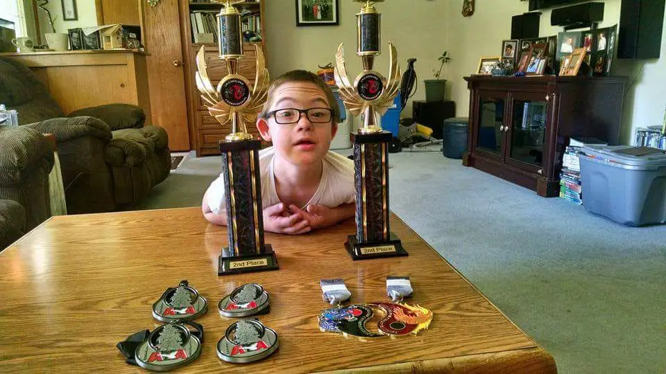 A person with glasses looks at the camera between two trophies, with medals on the table in front.