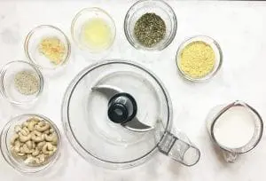 Raw cashews, almond milk, garlic, lemon juice, nutritional yeast, herbs, salt, pepper, and a food processor on a white surface.