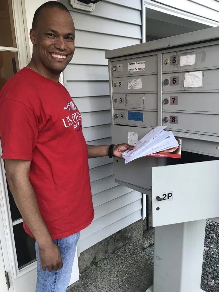 Dudley in a red t-shirt and jeans stands by a mailbox.