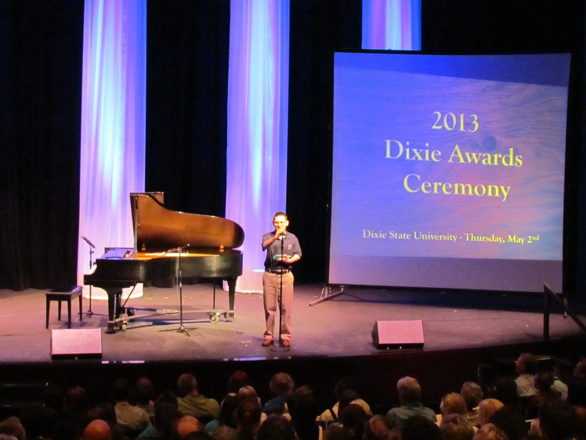 A man speaks at a podium on a stage with a piano and a large screen displaying "2013 Dixie Awards Ceremony".