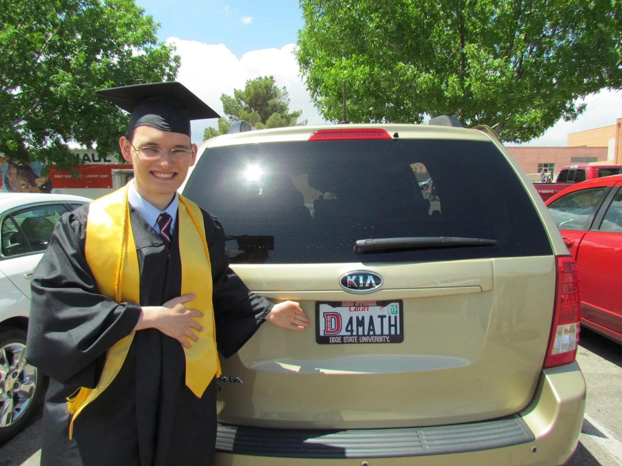 Joshua in graduation cap and gown points to a license plate that reads "I 4 MATH".