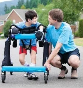 A man in a blue shirt crouches beside a young boy using a walker.