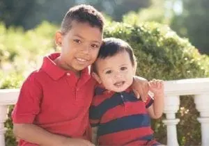 Two young brothers, one older with dark hair and a red polo shirt, the other a baby in a striped shirt, smile at the camera.