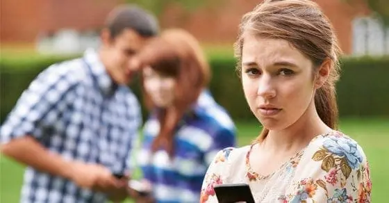 A young girl looks worriedly at her phone while two teenagers whisper behind her.