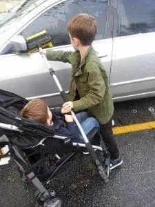 A young boy cleans a car window while another boy sits in a stroller beside him.