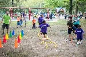 Children and adults participate in an outdoor obstacle course with cones and hurdles.