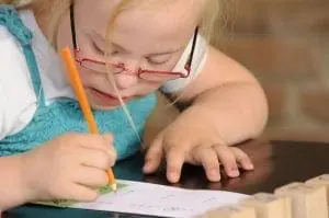 Young child with Down syndrome wearing glasses and writing with a pencil.